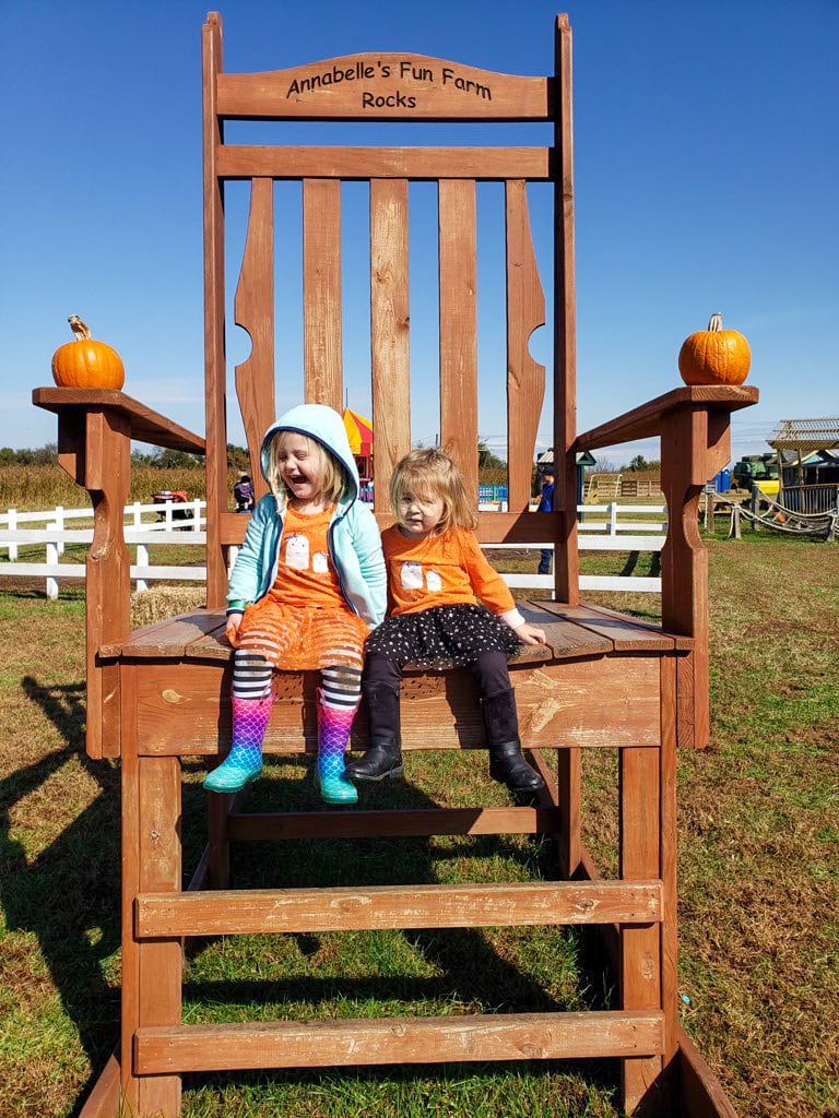 wo young girls sit on an oversized wooden chair with "Annabelle's Fun Farm Rocks" carved into the top. Each armrest is decorated with a pumpkin, and the girls wear festive orange outfits with fall patterns. The giant chair makes them look tiny, creating a playful photo opportunity at the farm.