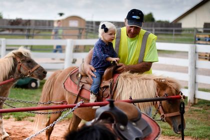 A young girl with a navy bow in her hair sits on a small pony while an adult in a neon safety vest helps guide her at Chester’s Party Barn & Farm. The pony ride is set within a fenced area, creating a safe and fun hands-on animal experience for kids.