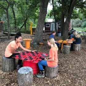 Two girls sit on tree stump seats playing checkers on a red and black board set atop a barrel in an outdoor area covered with wood chips. Behind them, other visitors sit at picnic-style tables beneath tall trees. The shaded setup creates a cozy, rustic play space at the farm.