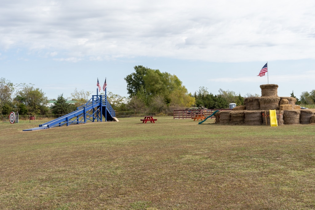 An open grassy field features a large blue wooden slide with American flags at the top and a hay bale play structure stacked in tiers with a yellow slide, also topped with a flag. Picnic tables are scattered around the area, and an archery target sits off to the side. The setup creates a playful farm-themed activity space.