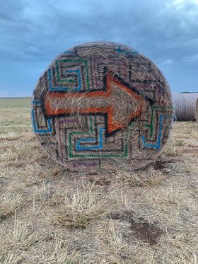 A large round hay bale at Jahn’s Pumpkin Hill is painted with a colorful maze design and a bold orange arrow pointing right. Set against a wide-open field and cloudy sky, the hay bale serves as a creative and fun directional marker for visitors exploring the farm.