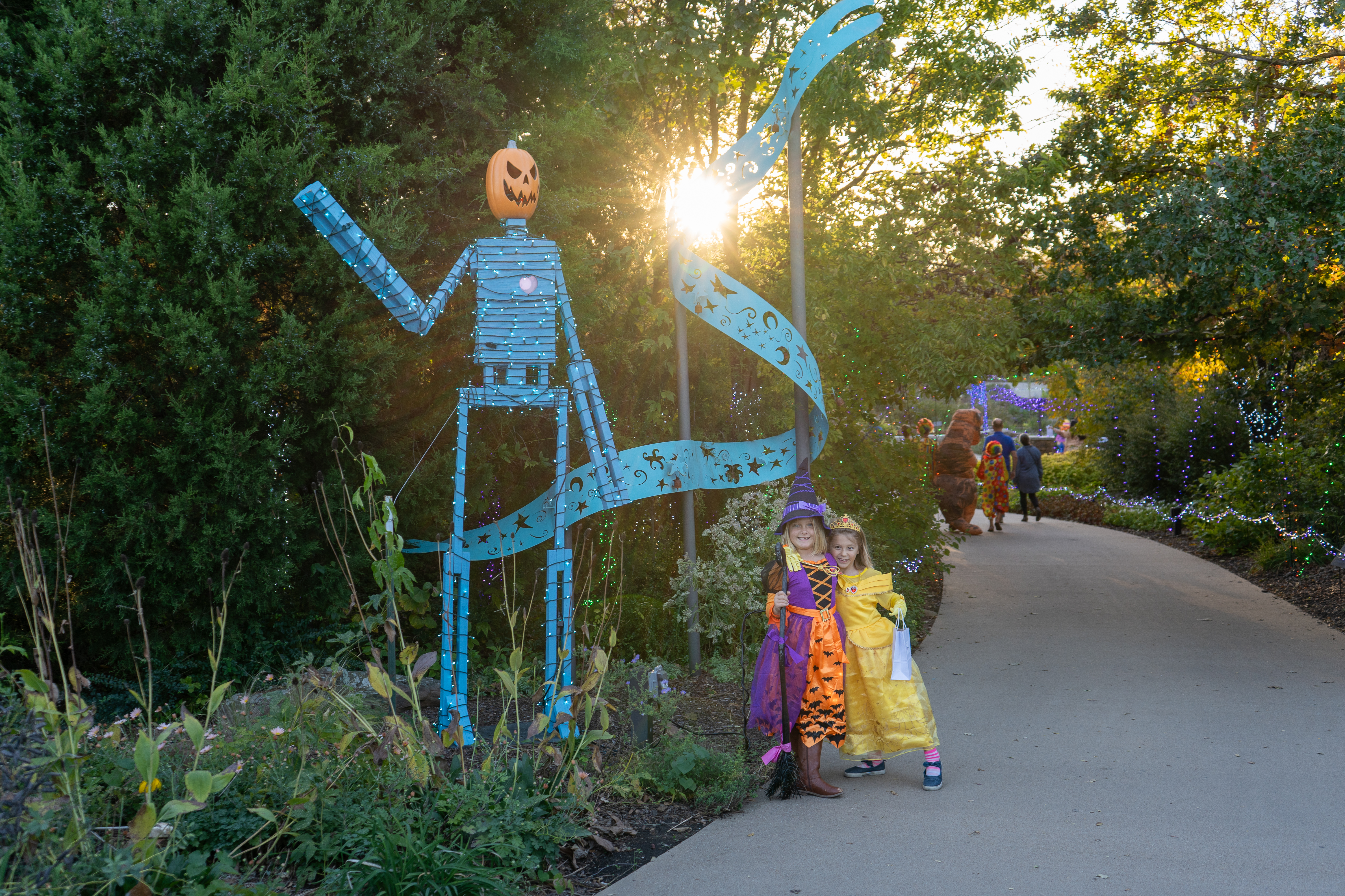 Tulsa Botanic Garden - Two children dressed in Halloween costumes, one as a witch and the other as Belle from Beauty and the Beast, pose on a garden pathway. Beside them stands a tall blue metal figure with a pumpkin head, lit by the setting sun behind it. The path is lined with greenery and festive lights, creating a whimsical fall scene.