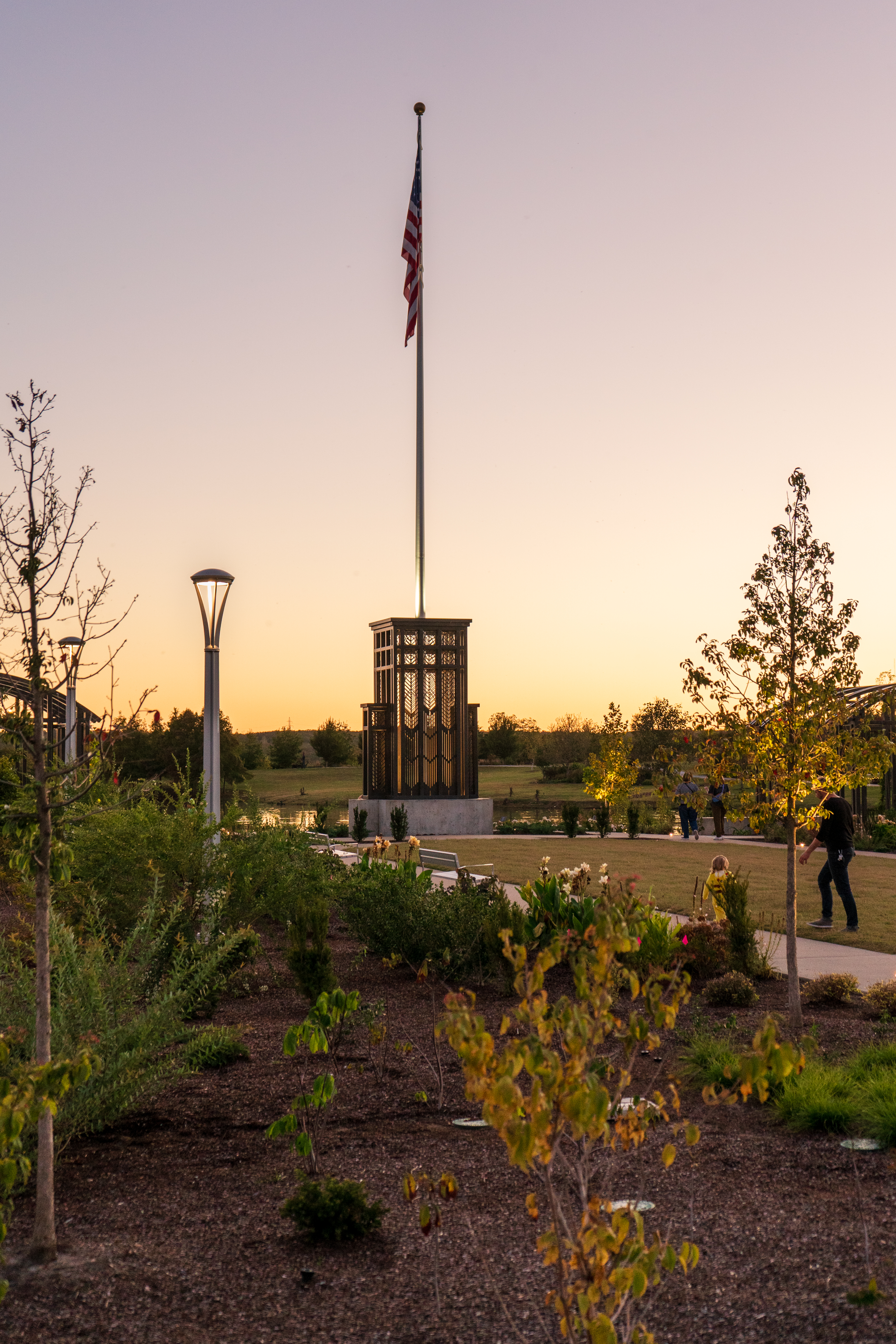 Tulsa Botanic Garden - A tall American flag waves atop a flagpole at sunset, rising above a decorative metal monument surrounded by landscaped gardens. Soft golden light fills the sky, casting a warm glow on the trees and walking path below. People stroll nearby, enjoying the peaceful evening scene.