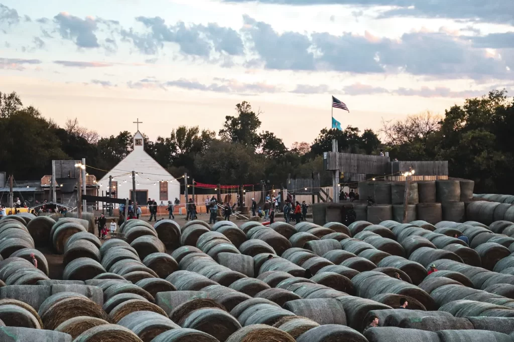 Dozens of large round hay bales are stacked to form a winding hay maze at Brown Ranch, with families exploring the paths as the sun sets. In the background, a white chapel-style building and American flag add charm to the festive fall setting.