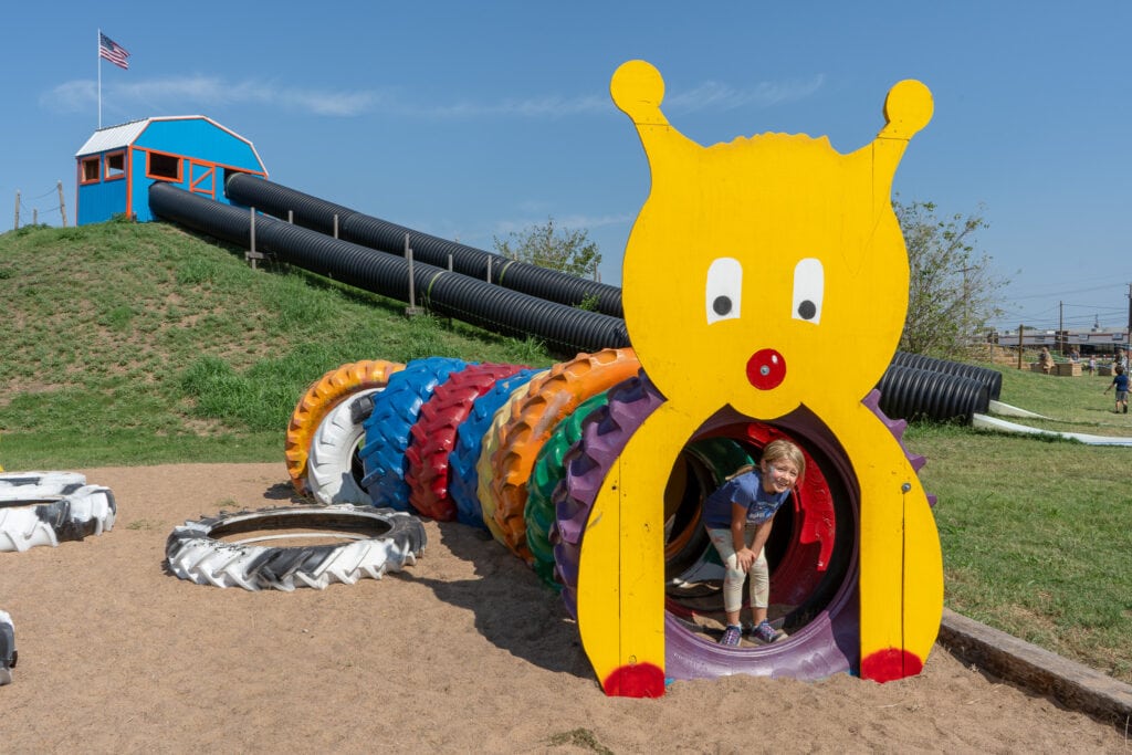 A child crawls through a colorful caterpillar tunnel made of painted tires with a large yellow cartoon caterpillar face at the entrance. Behind the playground, a tall slide built into a grassy hill leads down from a small blue and orange playhouse with an American flag on top. The bright colors and playful setup create a cheerful outdoor play area.