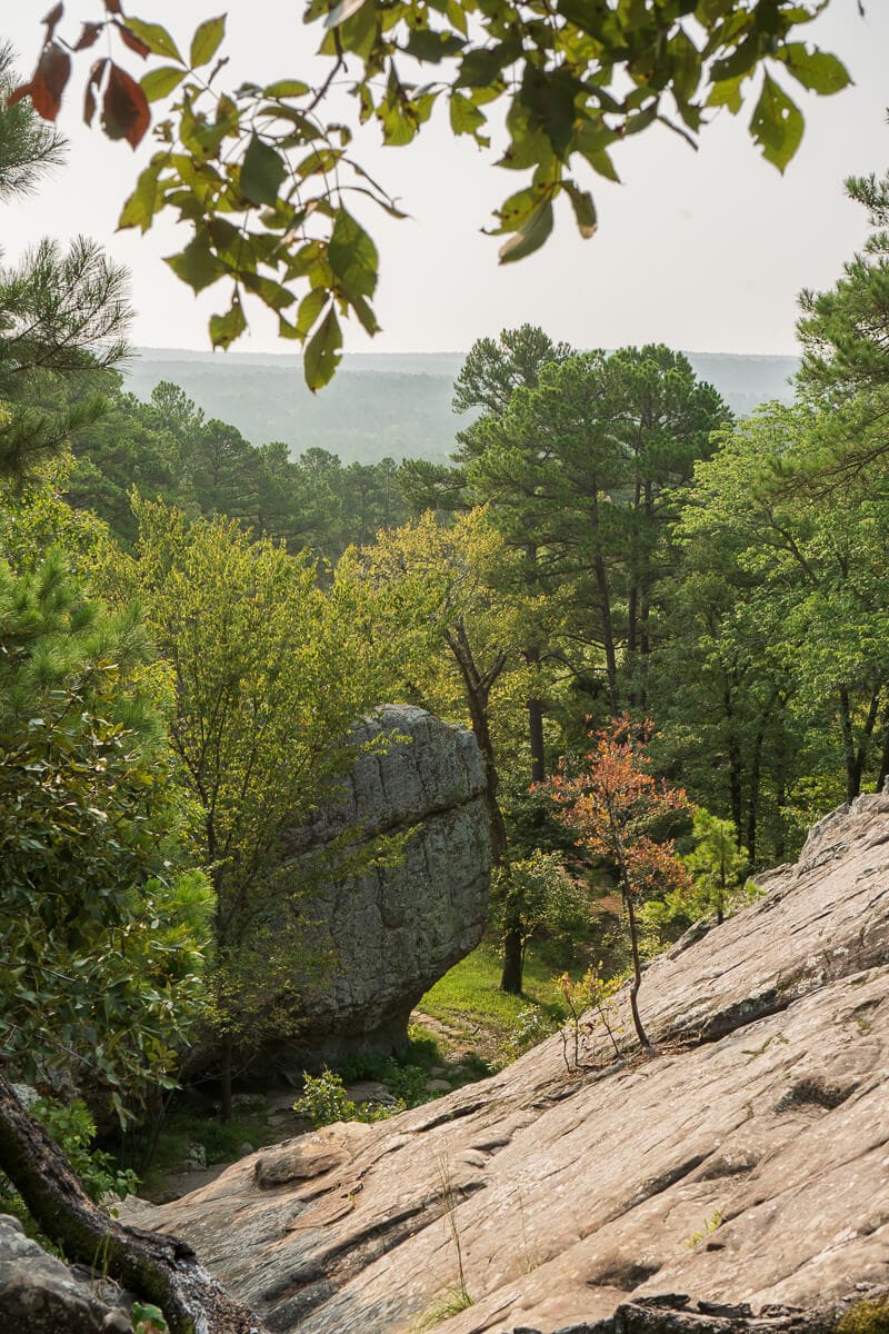 Large boulder nestled among dense green trees at Robbers Cave State Park, viewed from a sloped rock outcrop in the foreground. The scene is framed by leafy branches, with a hazy horizon visible in the distance.