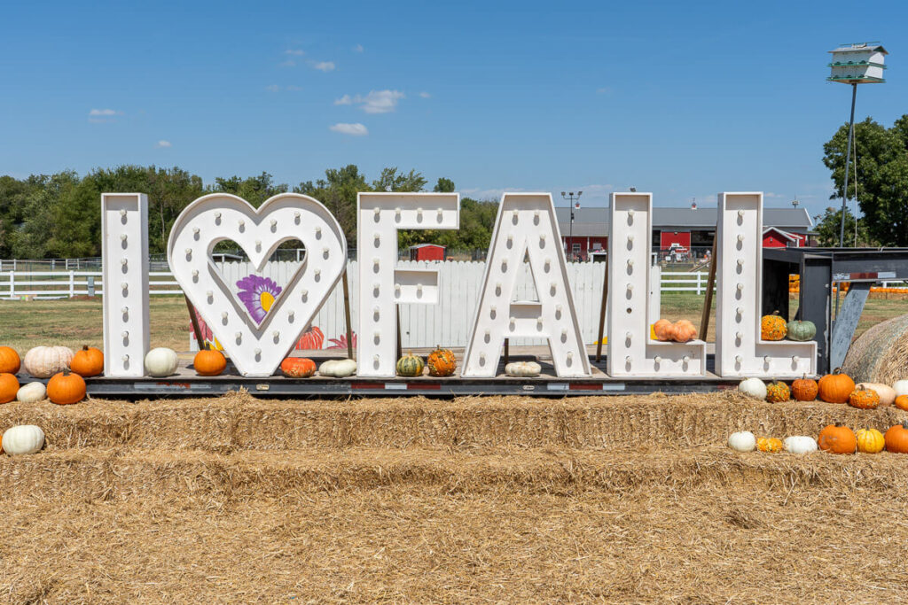 Large marquee letters spell out “I ♥ FALL” at Orr Family Farm, with a heart replacing the word "love" and lights outlining each letter. The sign is surrounded by colorful pumpkins and gourds on hay bales, creating a festive autumn display at a pumpkin patch near Oklahoma City.