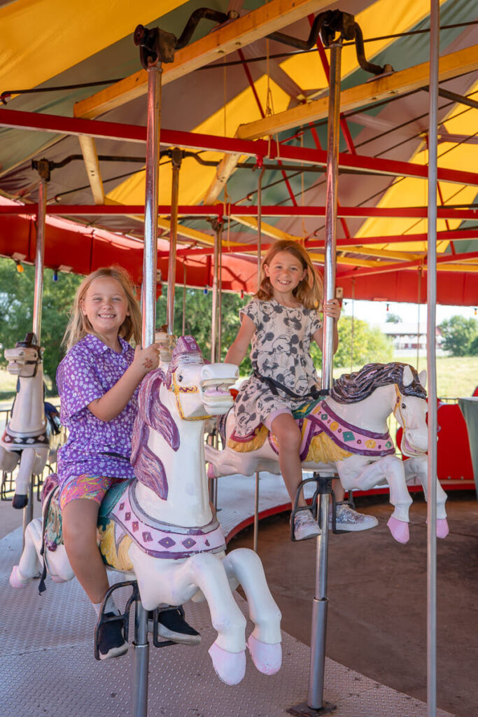 Two young girls smile while riding white carousel horses with purple and pink saddles under a bright red and yellow tent at Orr Family Farm. The colorful vintage carousel adds a whimsical, family-friendly charm to the Oklahoma City pumpkin patch experience.