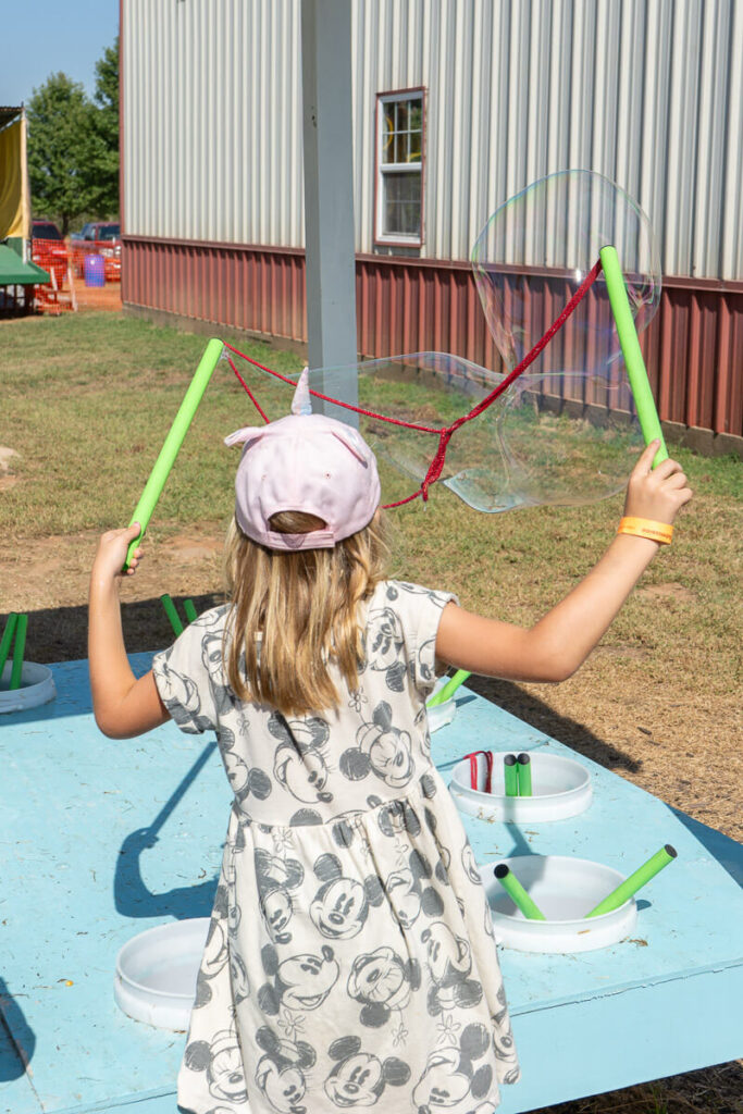 A young girl wearing a pink cap and a Mickey Mouse dress makes giant bubbles using green wands and a red string. She stands at a blue table with white trays of bubble solution in front of a barn-like building. A large bubble floats in the air beside her.