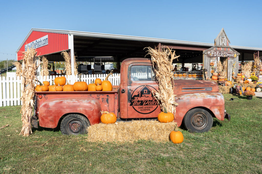An old red pickup truck decorated with pumpkins and cornstalks sits in front of a barn at a pumpkin patch. The truck bed is filled with pumpkins, and hay bales and more pumpkins surround it. A sign on the truck reads "The Patch Sand Springs Glenpool," marking it as a festive fall photo spot.