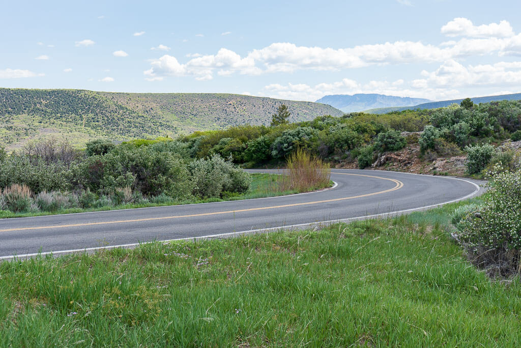 A winding two-lane road curves through lush greenery along the South Rim Drive at Black Canyon of the Gunnison National Park. The scenic route is surrounded by dense shrubs, rolling hills, and distant mountains under a bright blue sky with fluffy white clouds. A peaceful and picturesque drive showcasing the park's natural beauty.