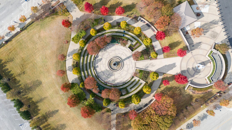 Aerial view of the John Hope Franklin Reconciliation Park in Tulsa, showcasing its circular plaza design centered around a prominent fountain. The park is surrounded by vibrant autumn trees in red, yellow, and green hues, with curving walkways and symmetrical landscaping leading to a secondary circular area with a statue.